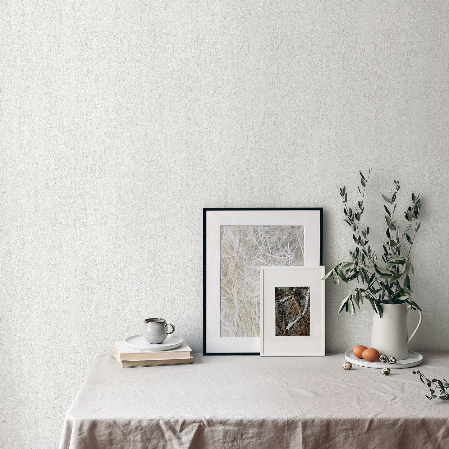 Neutral still life with two framed artworks, a white ceramic pitcher with greenery, a mug and books on beige linen table.