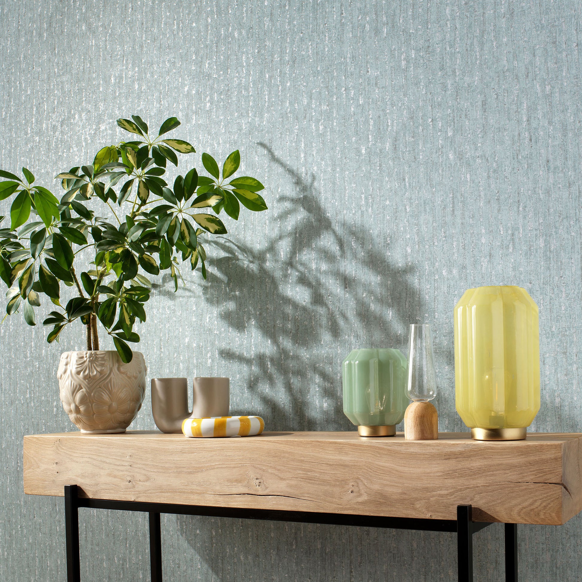 Wood console table with a potted plant, beige cups, mint-green lamp, clear vase, and tall yellow lamp against blue wall.