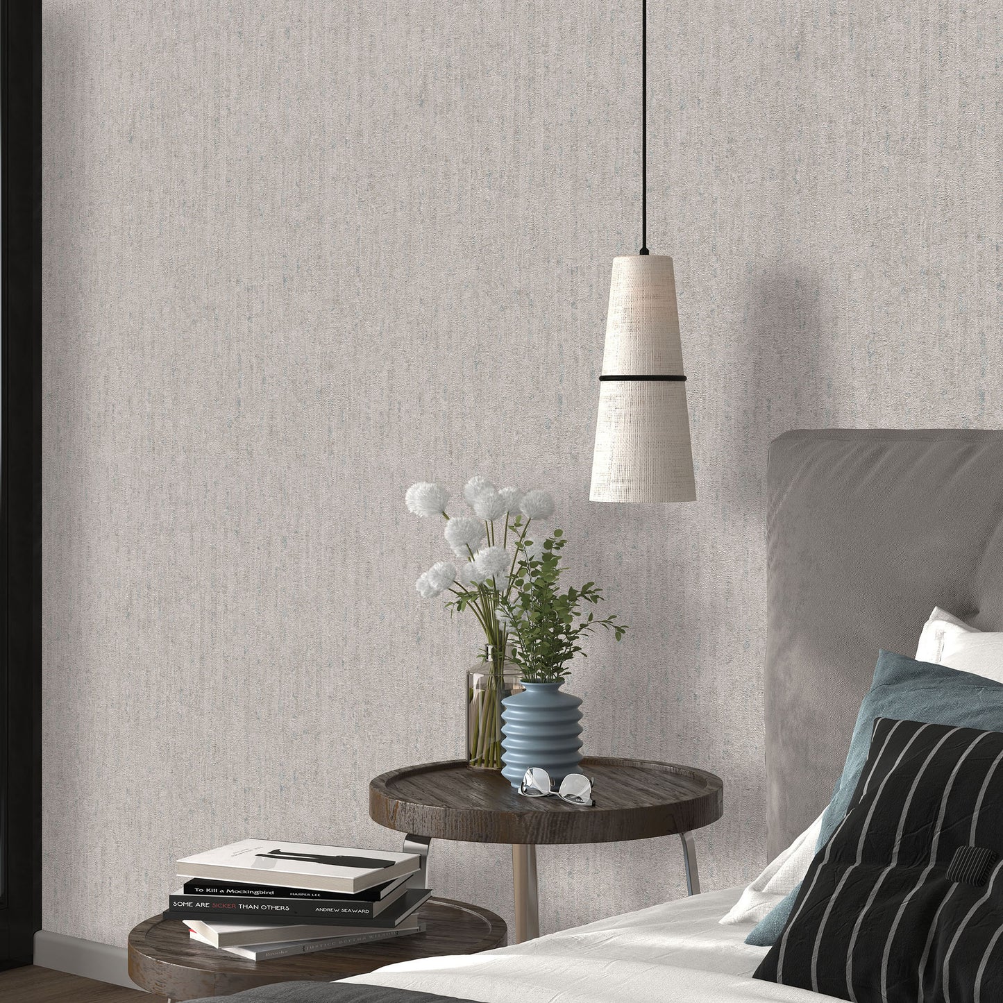 Taupe textured wallpaper with a traditional pattern visible in a bedroom scene; round wooden nightstand with a blue ribbed vase and white flowers, stacked books, gray upholstered bed, and a pendant light.