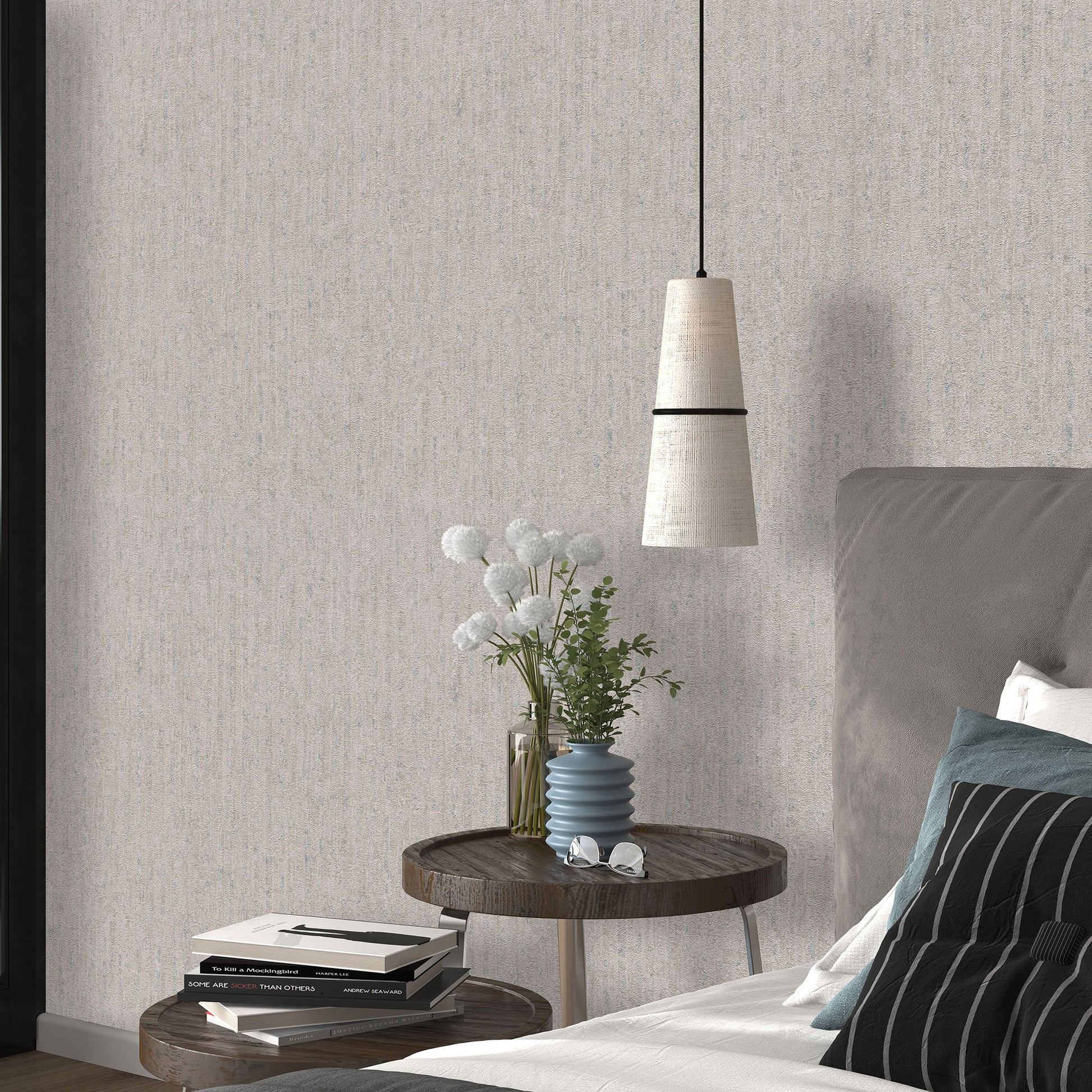 Taupe textured wallpaper with a traditional pattern visible in a bedroom scene; round wooden nightstand with a blue ribbed vase and white flowers, stacked books, gray upholstered bed, and a pendant light.