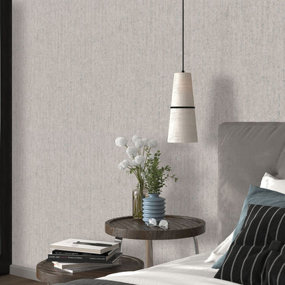 Taupe textured wallpaper with a traditional pattern visible in a bedroom scene; round wooden nightstand with a blue ribbed vase and white flowers, stacked books, gray upholstered bed, and a pendant light.