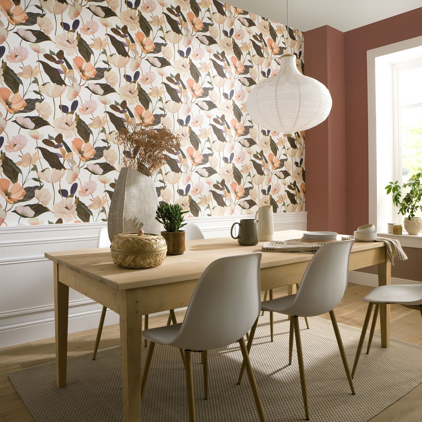 Light wood dining table with gray chairs, floral wallpaper in pinks and greens, wicker decor, and a round white pendant lamp.