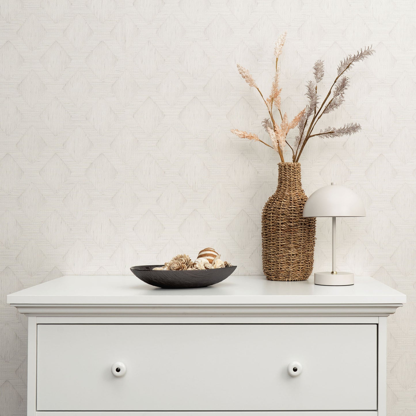 Gray wallpaper with a subtle pattern on the wall; foreground decor includes a white dresser, wicker vase with dried grasses, white dome lamp, and a black bowl with shells.