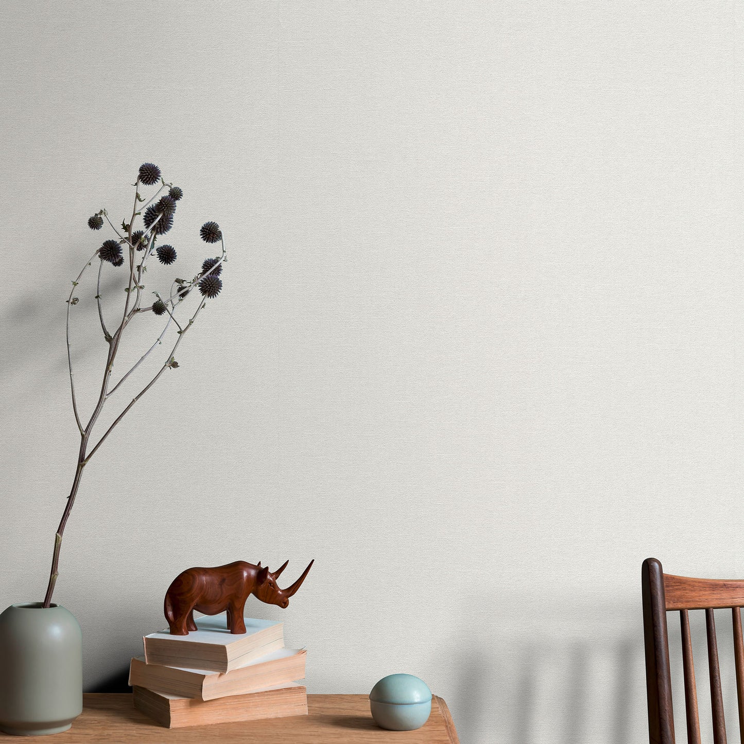Wallpaper background with minimalist desk decor: brown rhinoceros statue on beige books, pale grey-green vase with dried stems, teal ball, and a wooden chair.