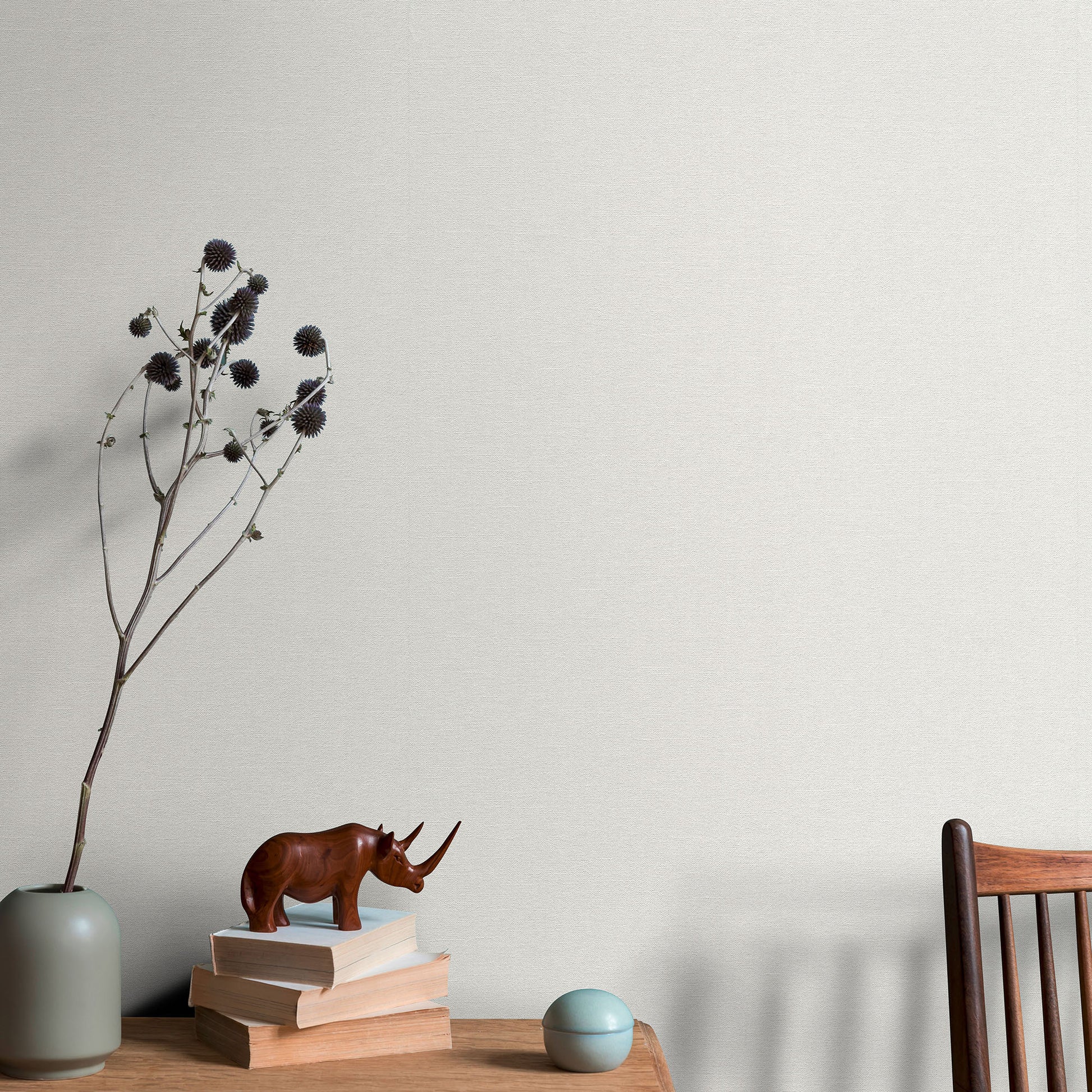Wallpaper background with minimalist desk decor: brown rhinoceros statue on beige books, pale grey-green vase with dried stems, teal ball, and a wooden chair.