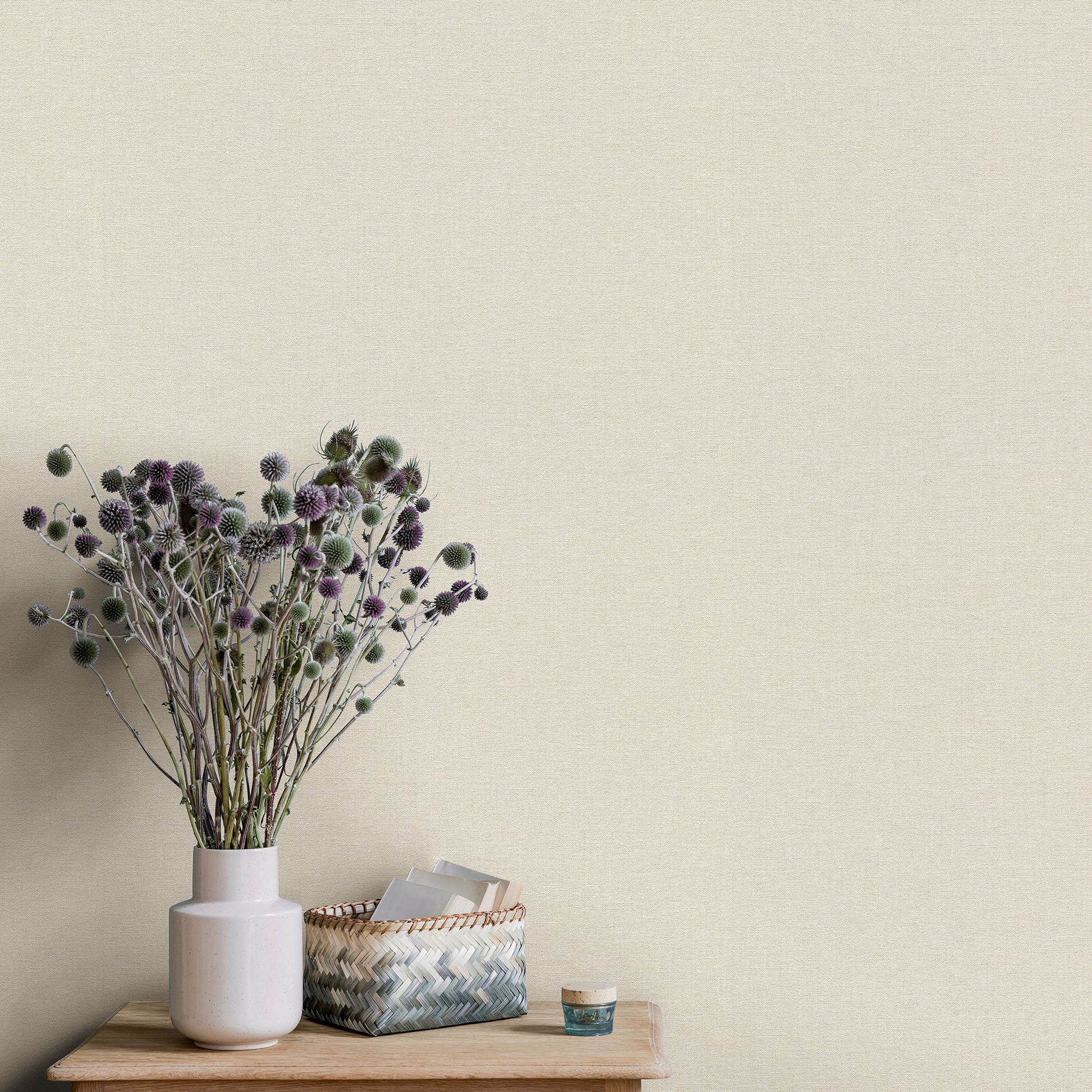 Beige textured wallpaper background with a pale pink ceramic vase of dried purple thistle stems on a wooden table, plus a woven basket and small jar.