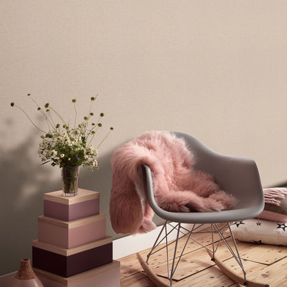 Pink linen-textured wallpaper visible on the walls in a room setting, with a gray molded chair with metal legs, a pink faux fur throw, pastel stacked boxes used as a side table, and a vase of white flowers.