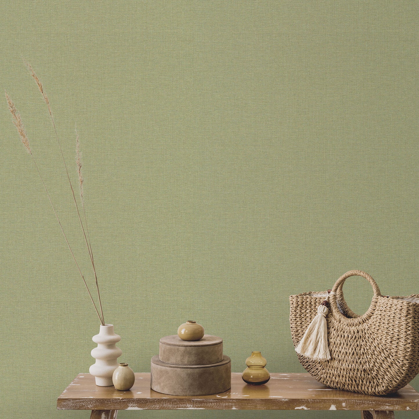 Olive green textured wall with wooden table holding nested boxes, ceramic vases with dried grasses, and woven straw handbag.