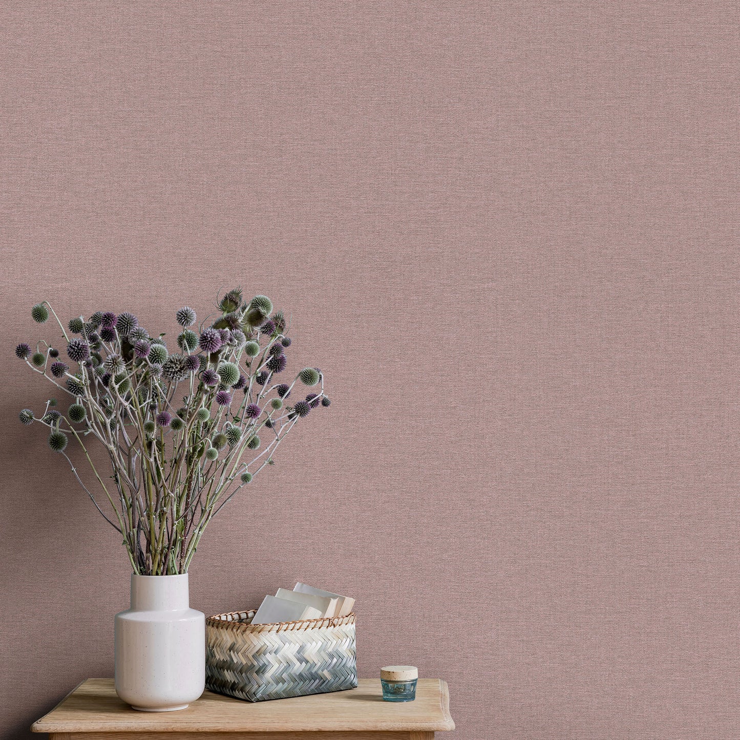 Soft pink textured wallpaper with a white vase of dried purple-topped stems on a wooden table, plus a woven basket and a small jar.