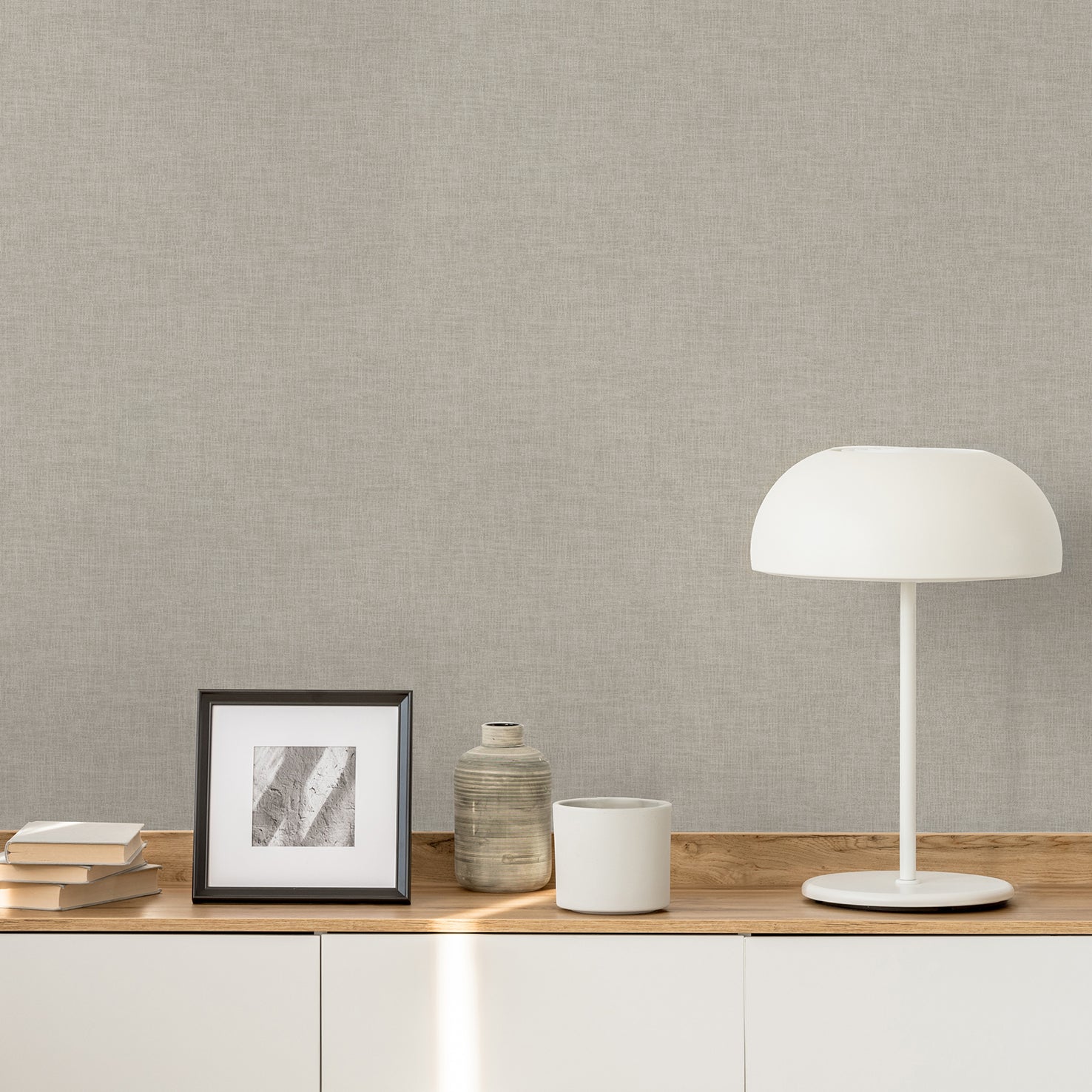 Taupe linen-weave textured wallpaper on a wall, with a wooden console, framed grayscale artwork, ceramic jar, white mug, books, and a white dome lamp.