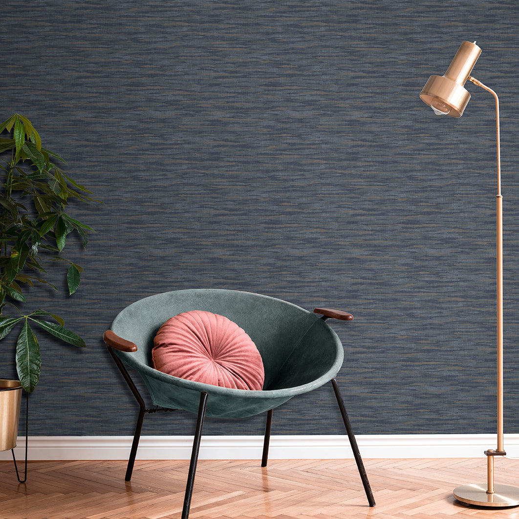 Teal velvet accent chair with wooden arms and black legs, pink round cushion; copper floor lamp, green plant, textured wall.