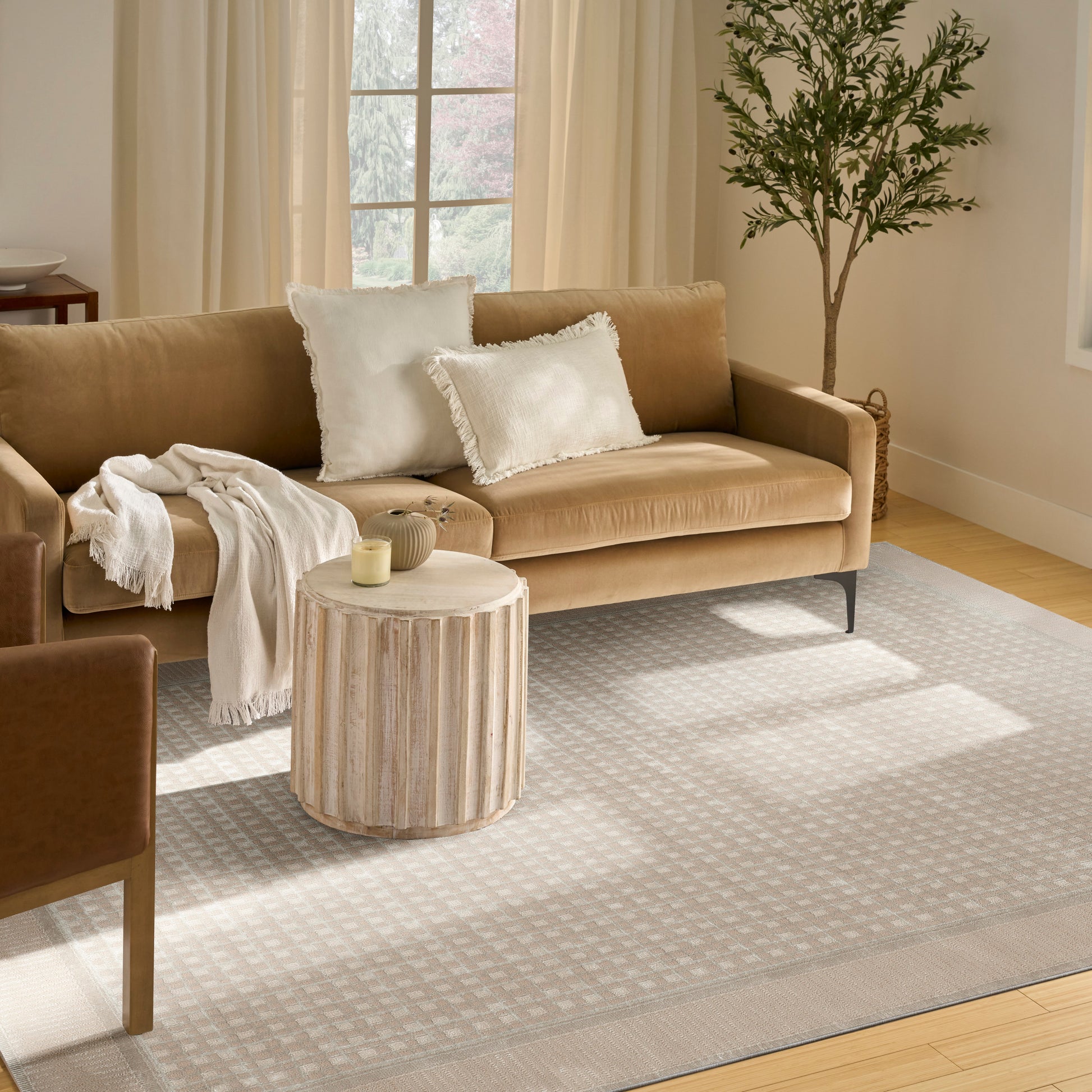 Beige sofa with white fringed pillows, round grooved wood coffee table, gray geometric rug, cream curtains and a potted tree.