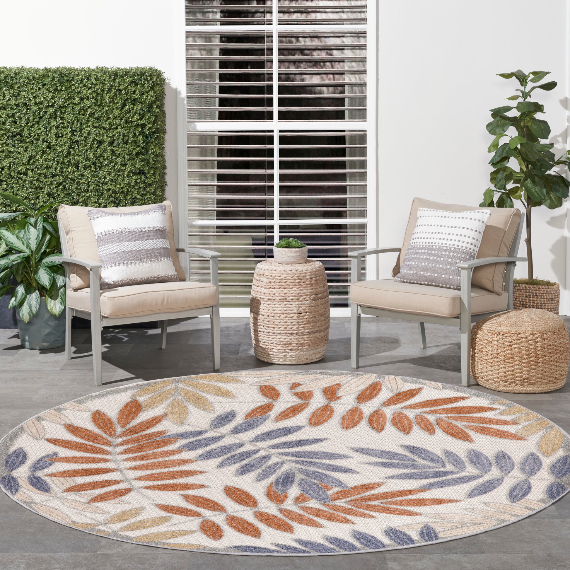 Round outdoor rug with orange, purple, beige leaf pattern on patio, flanked by beige cushioned metal chairs and woven stools.
