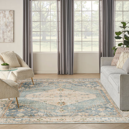 Bright living room with large window, gray drapes, beige armchair, light sofa, blue-cream patterned rug, glass side table.