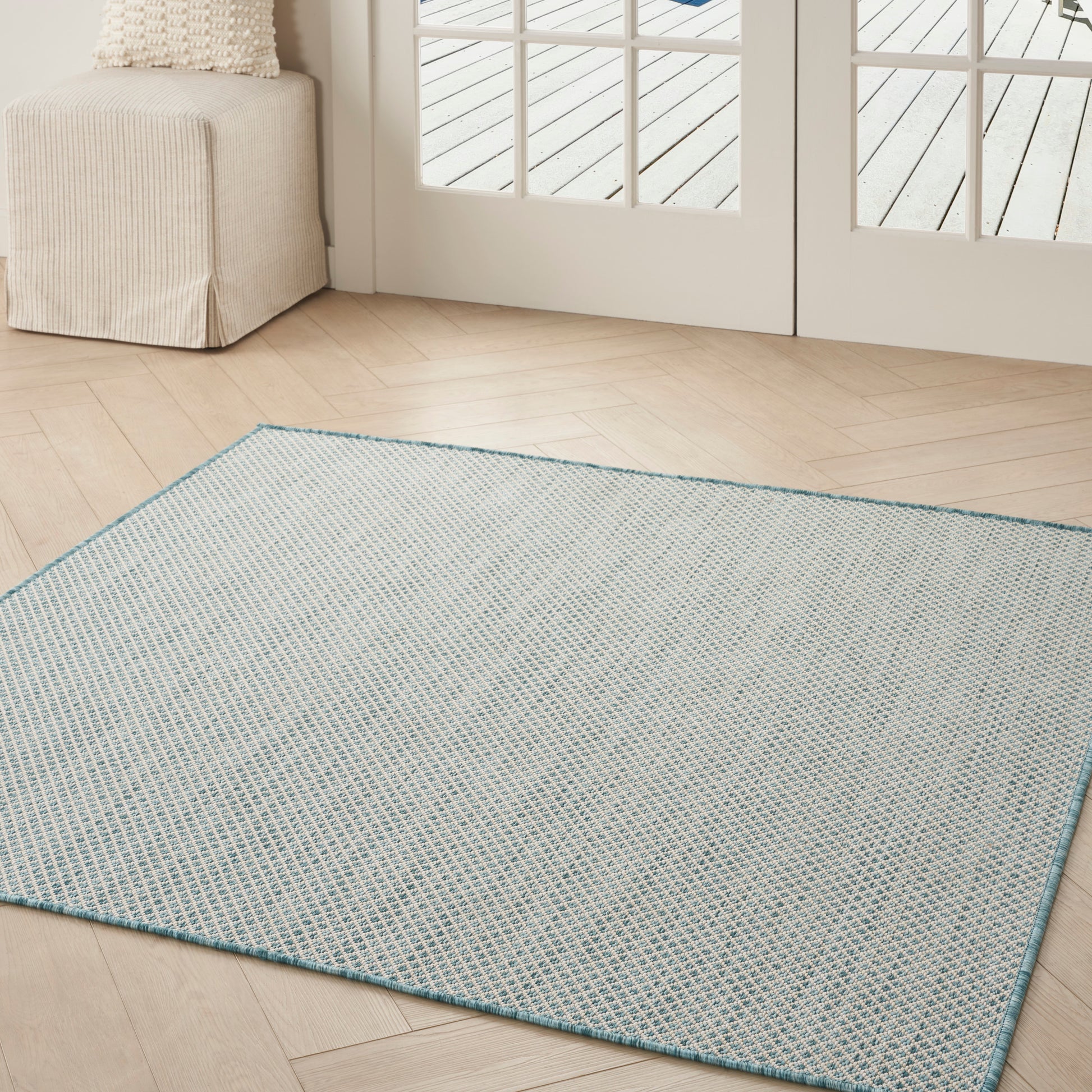 Square light blue woven area rug with teal border on light wood floor, beige ottoman and glass doors in a living room.