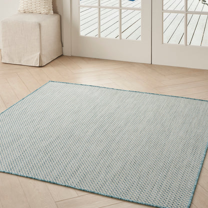 Square light blue woven area rug with teal border on light wood floor, beige ottoman and glass doors in a living room.