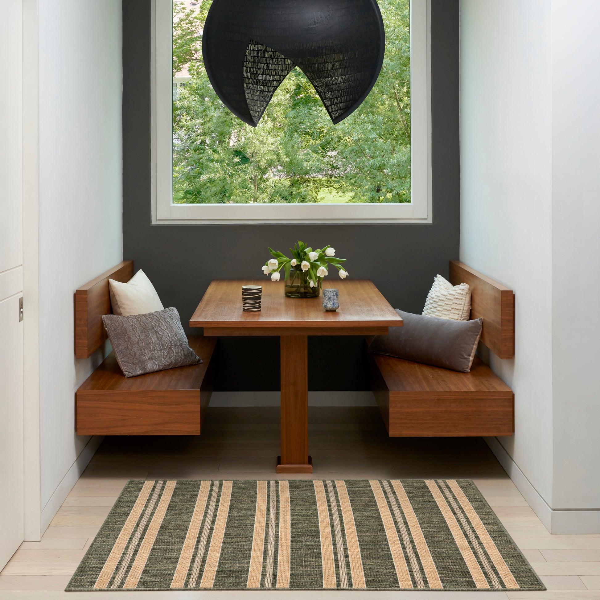 Corner dining nook with solid wood benches and table, striped beige-gray rug, black pendant light, window with greenery.