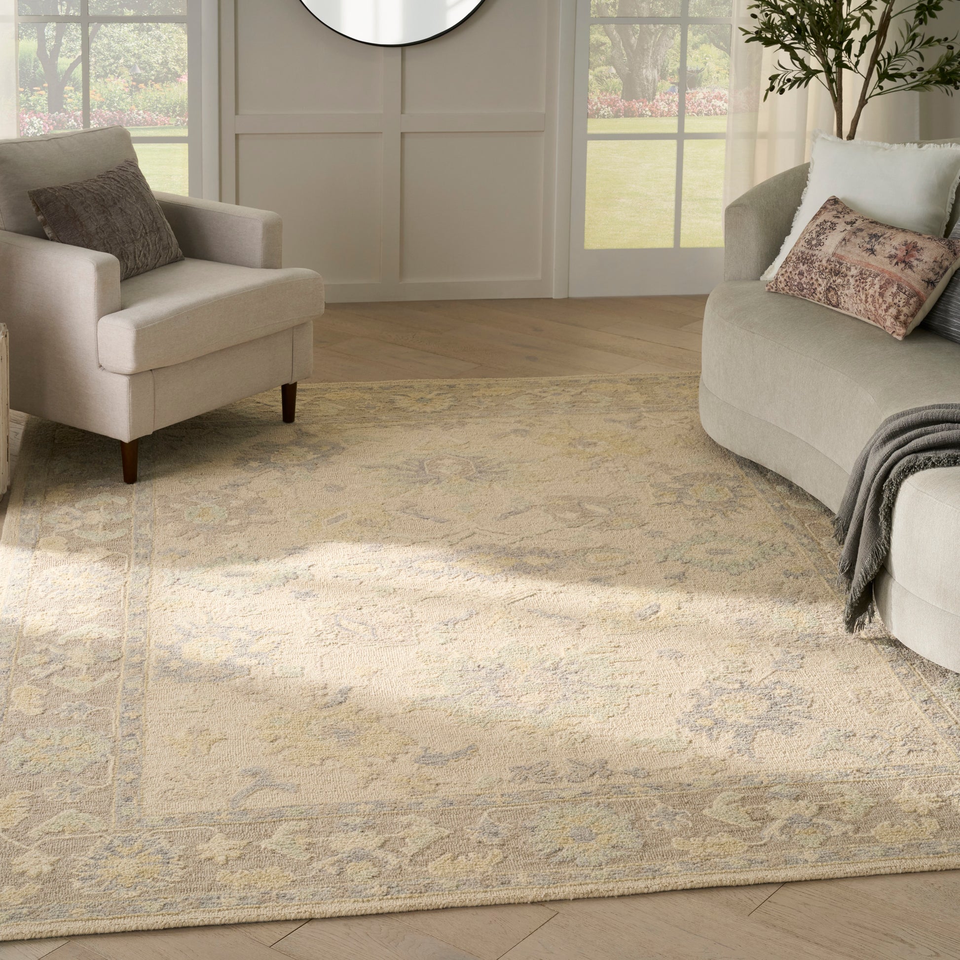 Neutral traditional area rug with ornate floral pattern in beige-gray tones, in a bright living room with sofa and armchair.
