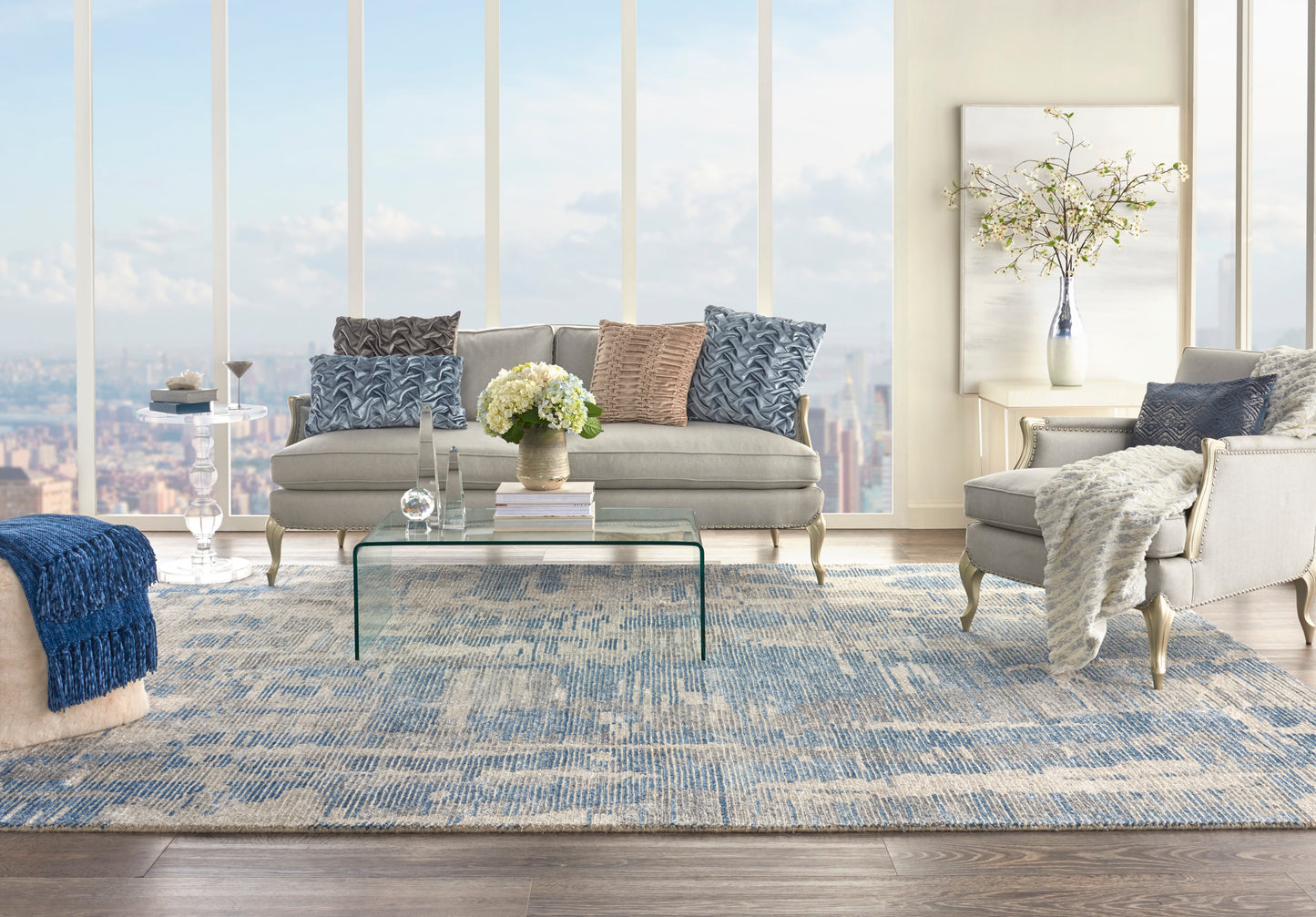 Light gray contemporary living room with blue cushions, glass coffee table, blue-beige rug, and city views.