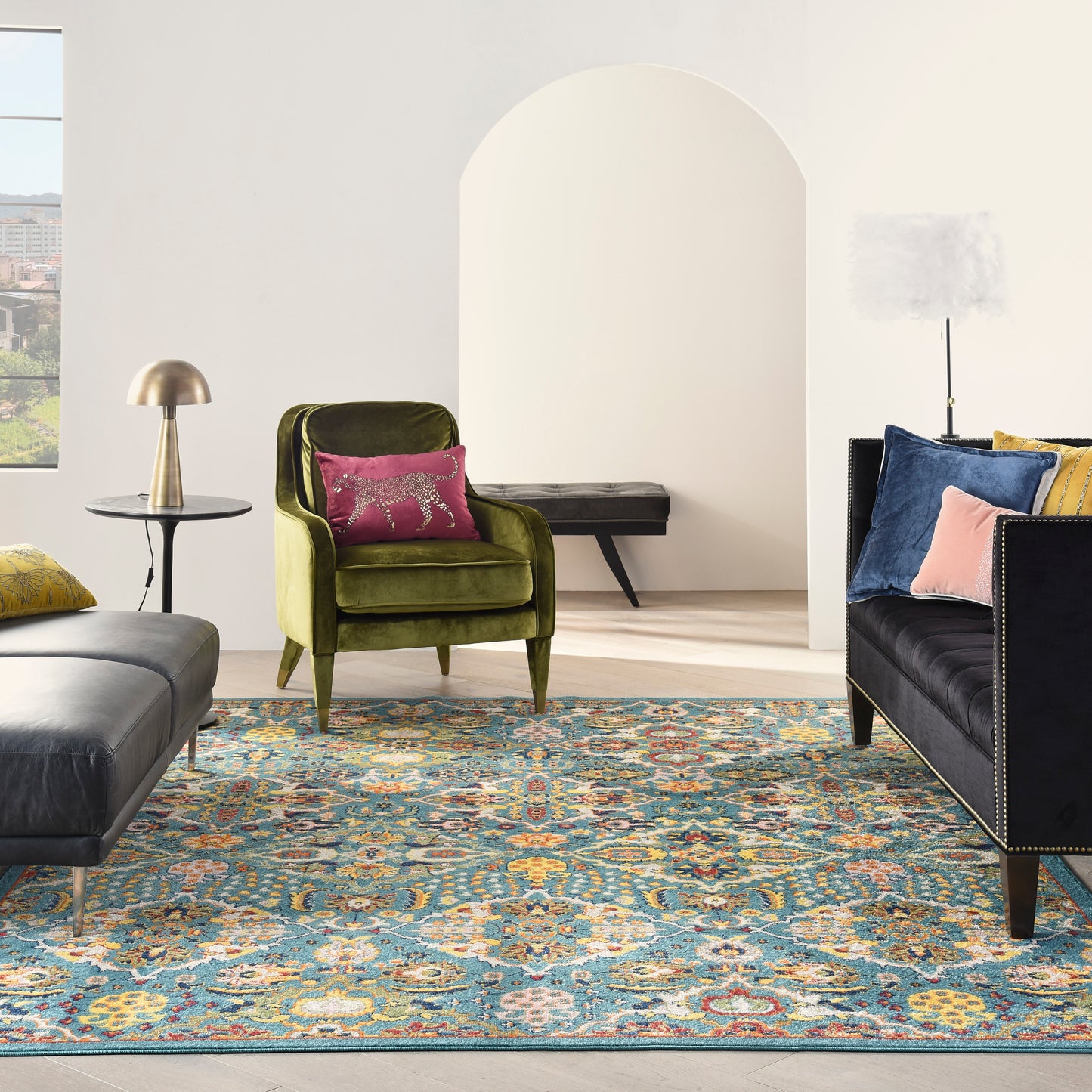 Olive green velvet armchair with pink pillow, dark tufted sofa, ornate blue-gold rug, brass lamp, light wood floor.