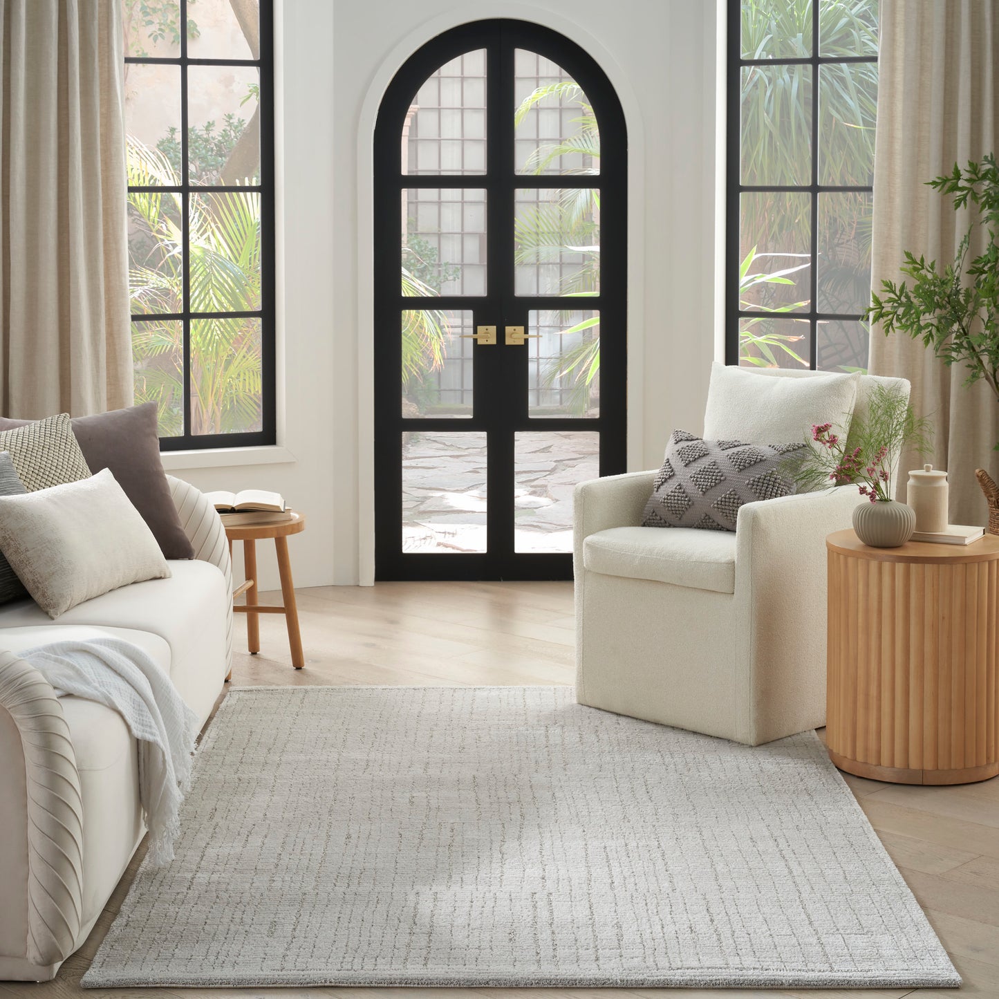 Light grey flatwoven area rug in a bright living room with cream sofa, neutral pillows, and an arched black door.