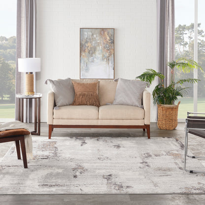 Beige mid-century sofa with gray and tan cushions, abstract wall art, side lamp, potted plant, and light gray patterned rug.