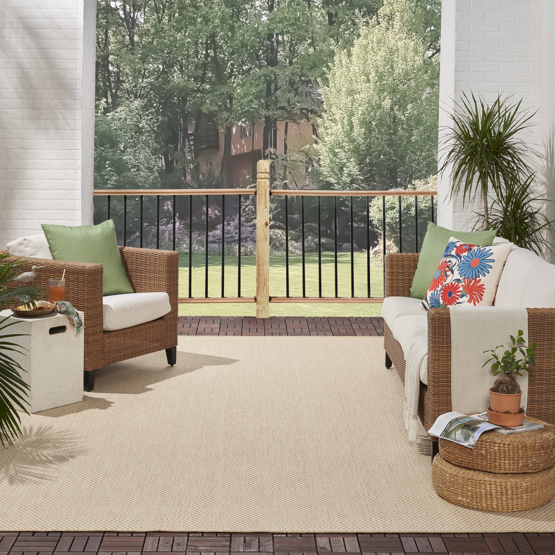 Wicker patio seating on a beige outdoor rug with white cushions, green pillows, a floral pillow, and potted plants.