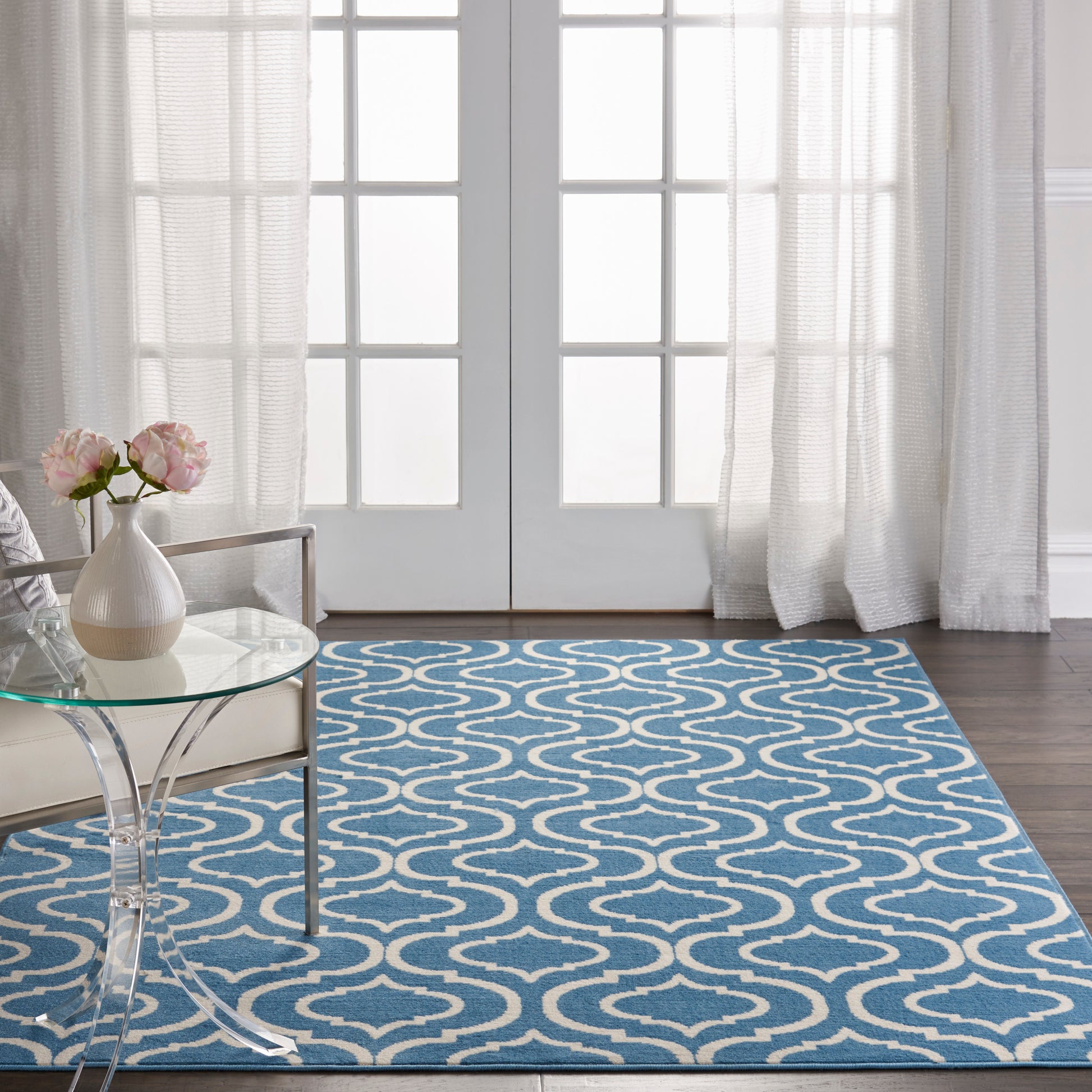 Blue area rug with white trellis pattern on dark wood floor, beside a clear glass coffee table and pink flowers.