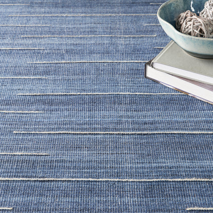 Blue woven rug with white horizontal stripes; stack of books topped by a teal bowl filled with twine.