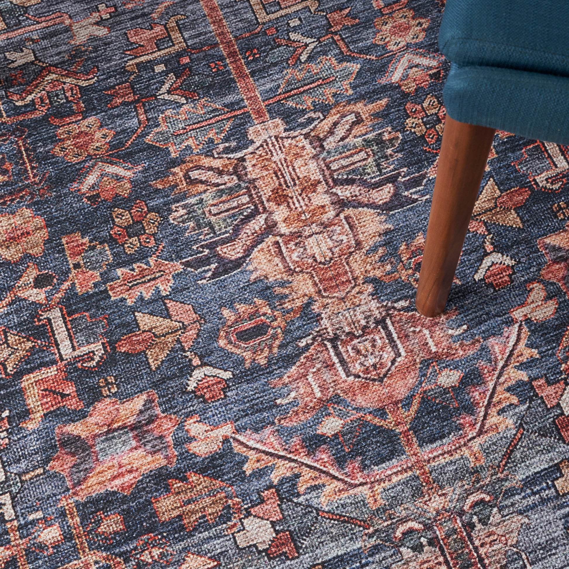 Blue oriental woven rug with pink, ivory & brown floral motifs; wooden chair leg resting on it.