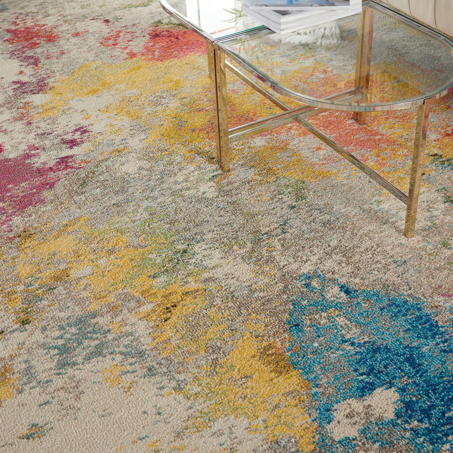 Colorful abstract rug with beige base and splashes of yellow, blue and pink; round glass coffee table with gold legs.