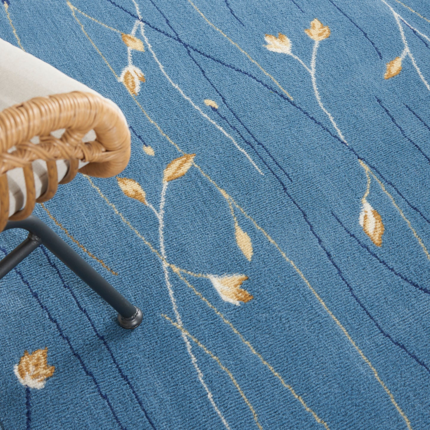 Blue rug with cream leaf motifs and white lines; woven texture beside a wicker chair with black metal legs.
