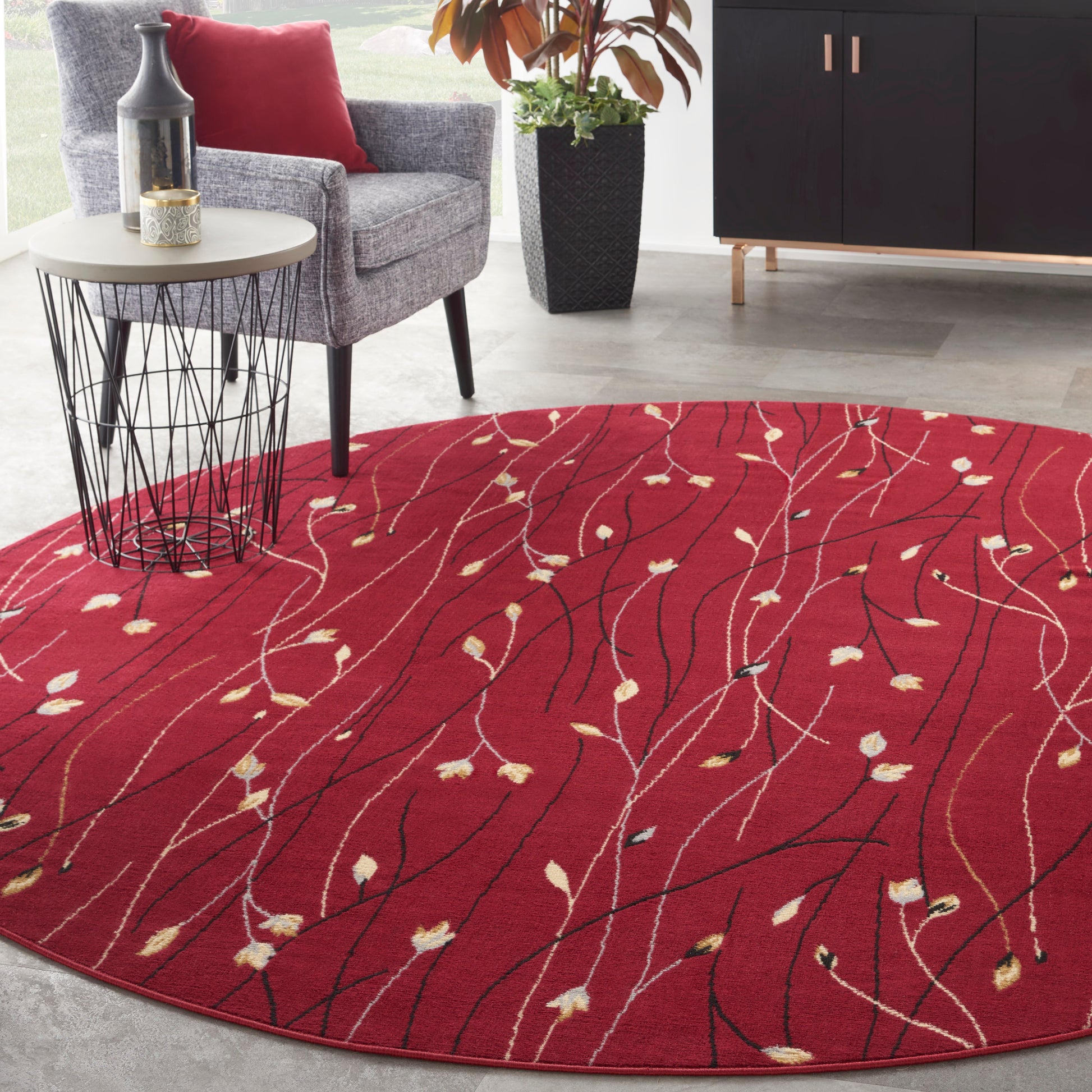 Red round area rug with white and beige leaf motifs and black lines; modern living room with grey chair and wire side table.