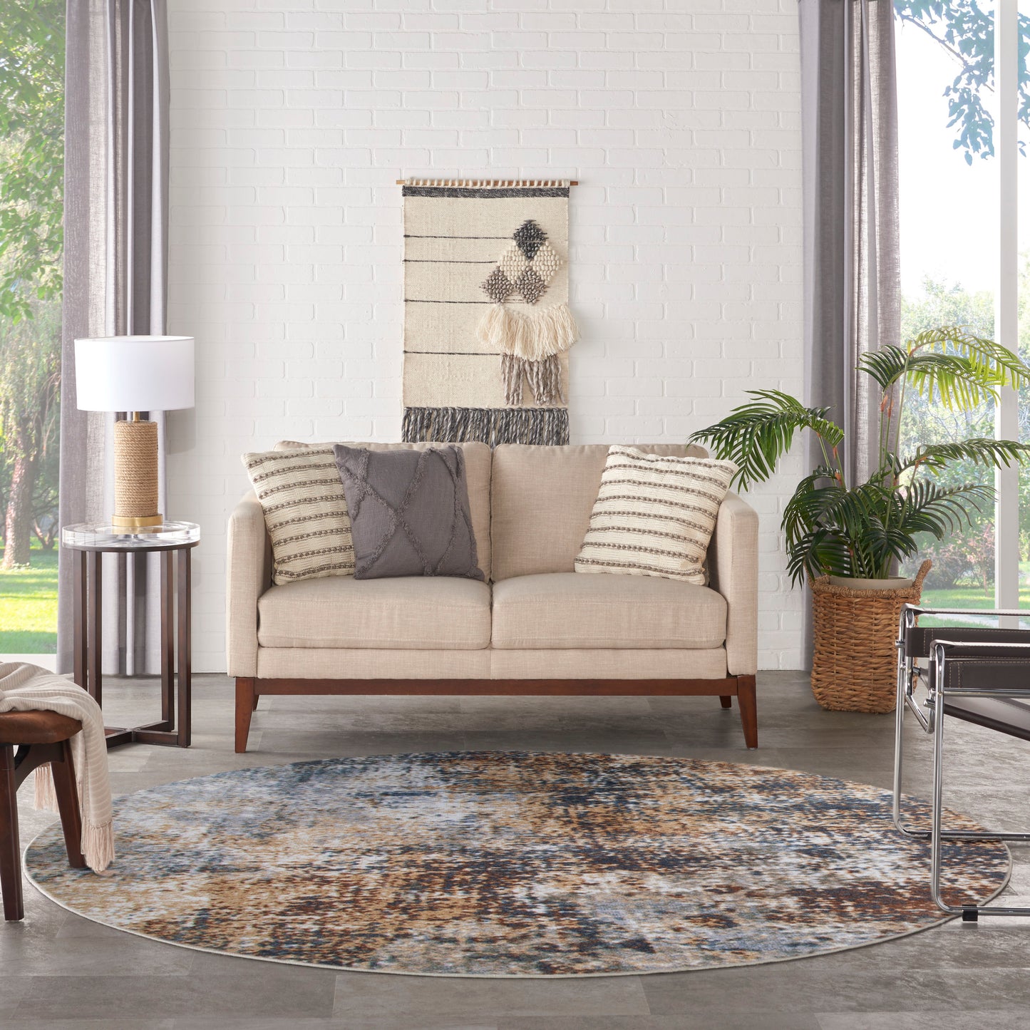Beige mid-century sofa with striped pillows in a bright living room; circular blue rug, woven wall hanging, plant, lamp.