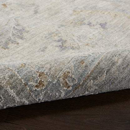 Light gray patterned area rug with muted blue and taupe accents, soft textured pile, rolled edge on dark wood floor.