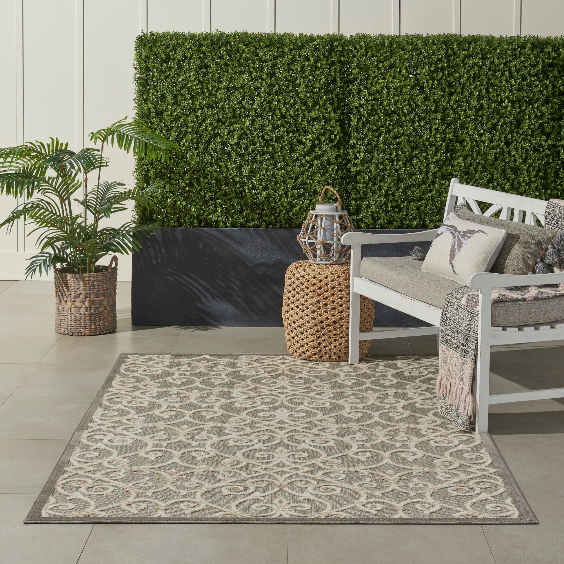 Beige taupe patterned outdoor rug on patio with white bench, cushions, wicker accents, lantern, potted plant and green hedge.