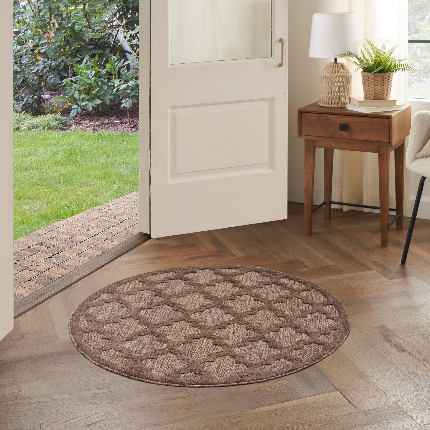 Round brown woven rug with a diamond pattern in an entryway on wood flooring, near a side table with a lamp.
