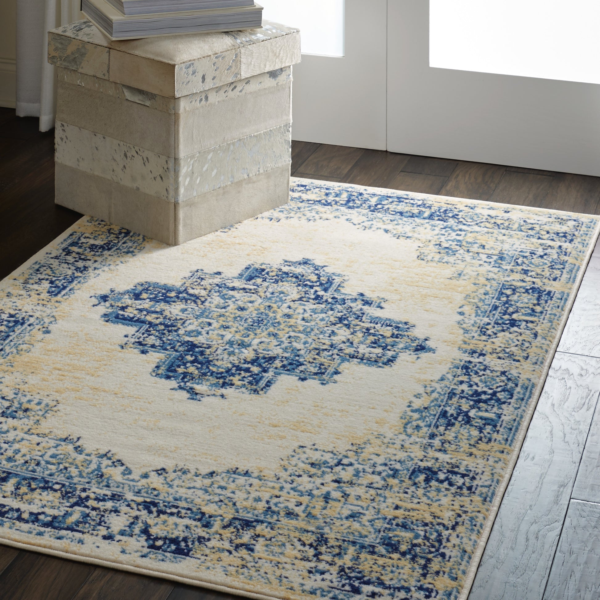 Cream & blue distressed area rug with medallion center on dark wood floor, beside beige stacked storage boxes & books.