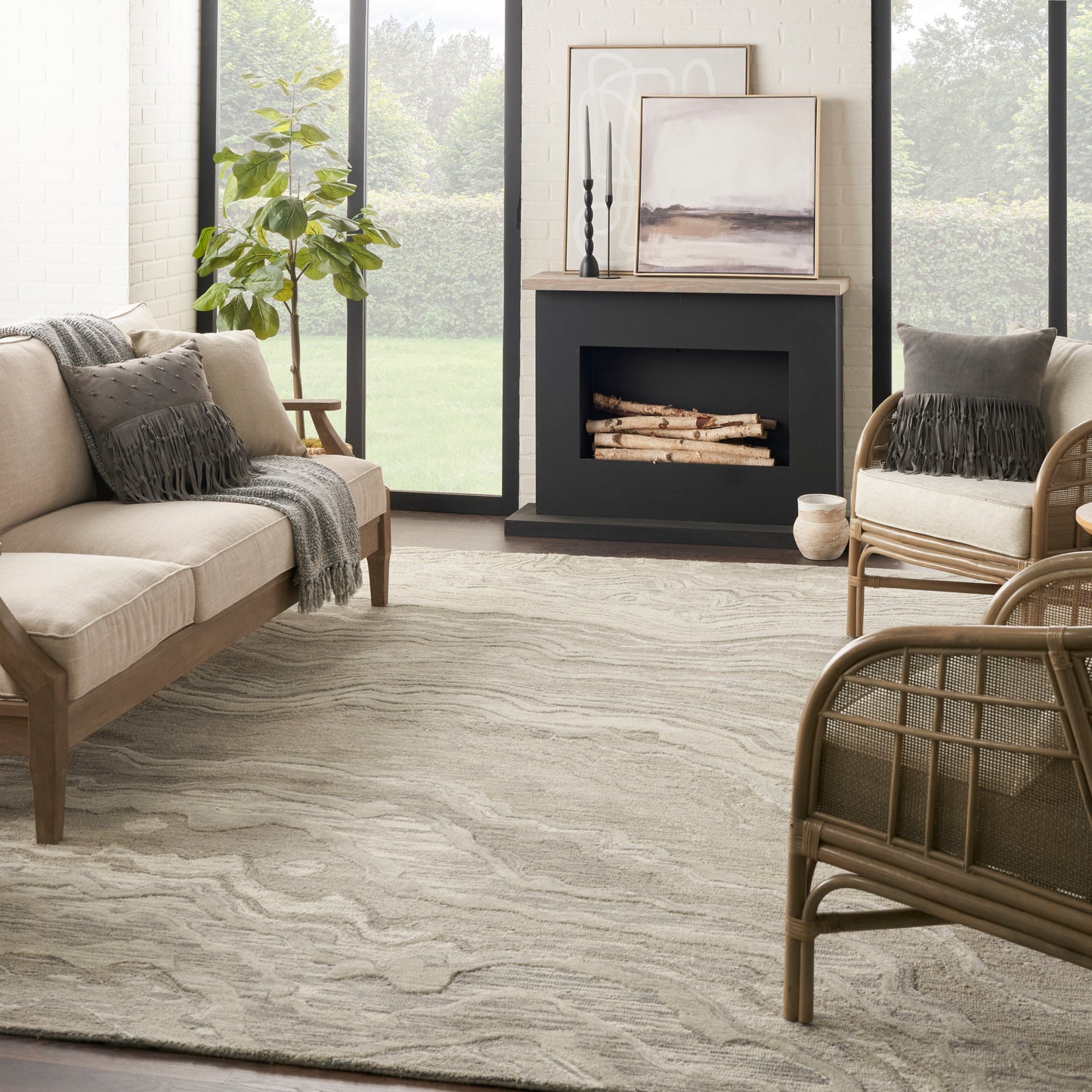 Neutral living room with beige fabric sofa, wicker chairs, light swirled rug, black fireplace, potted plant, and framed art.