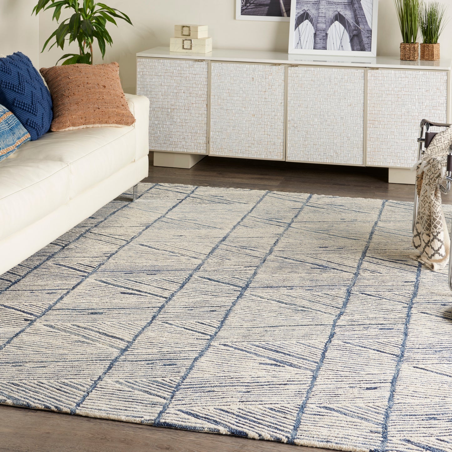 Cream sofa with blue and brown throw pillows, white woven sideboard, potted plant, and blue geometric grid rug.