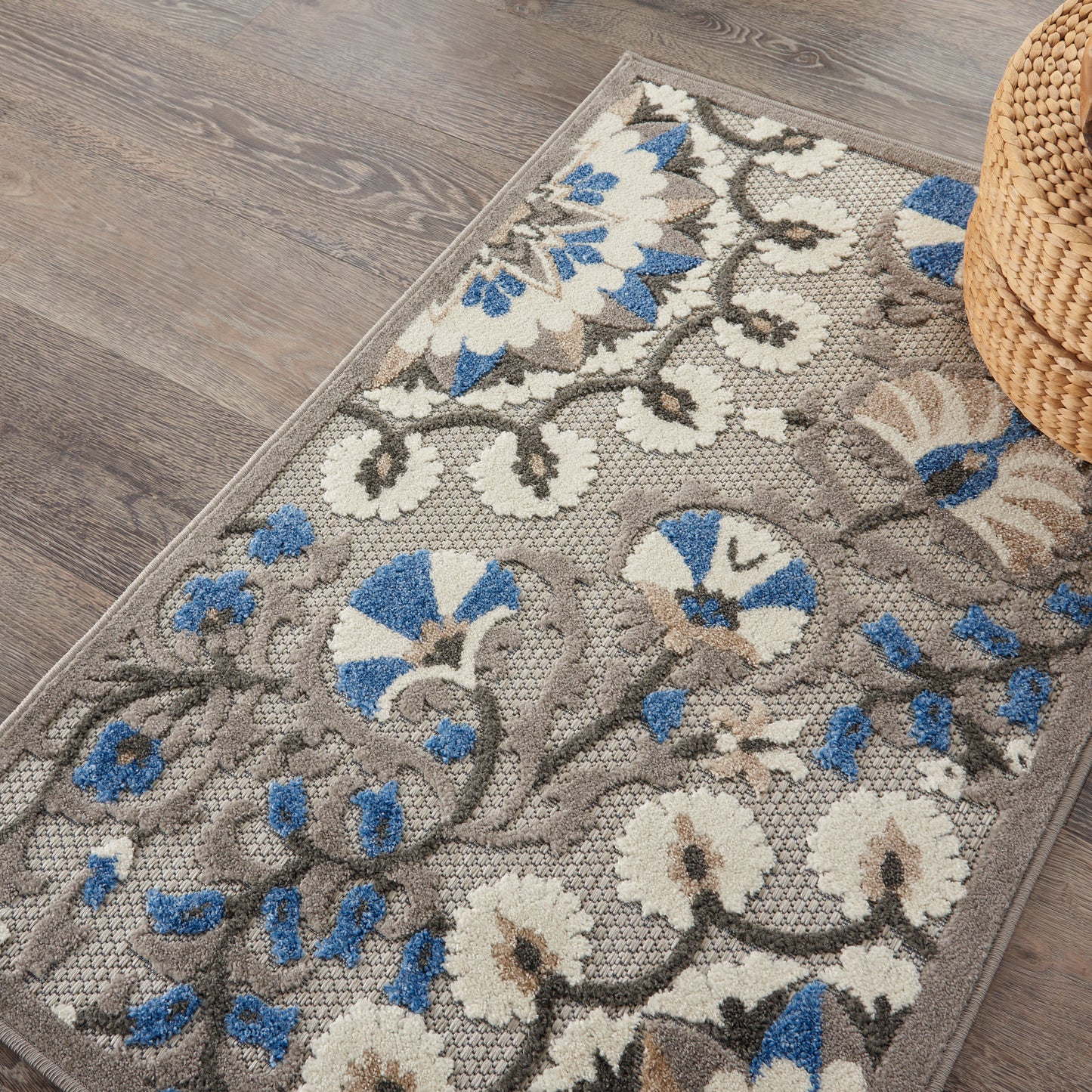 Gray floral rug with blue and ivory flowers, woven pattern, on a wood-look floor beside a woven basket.