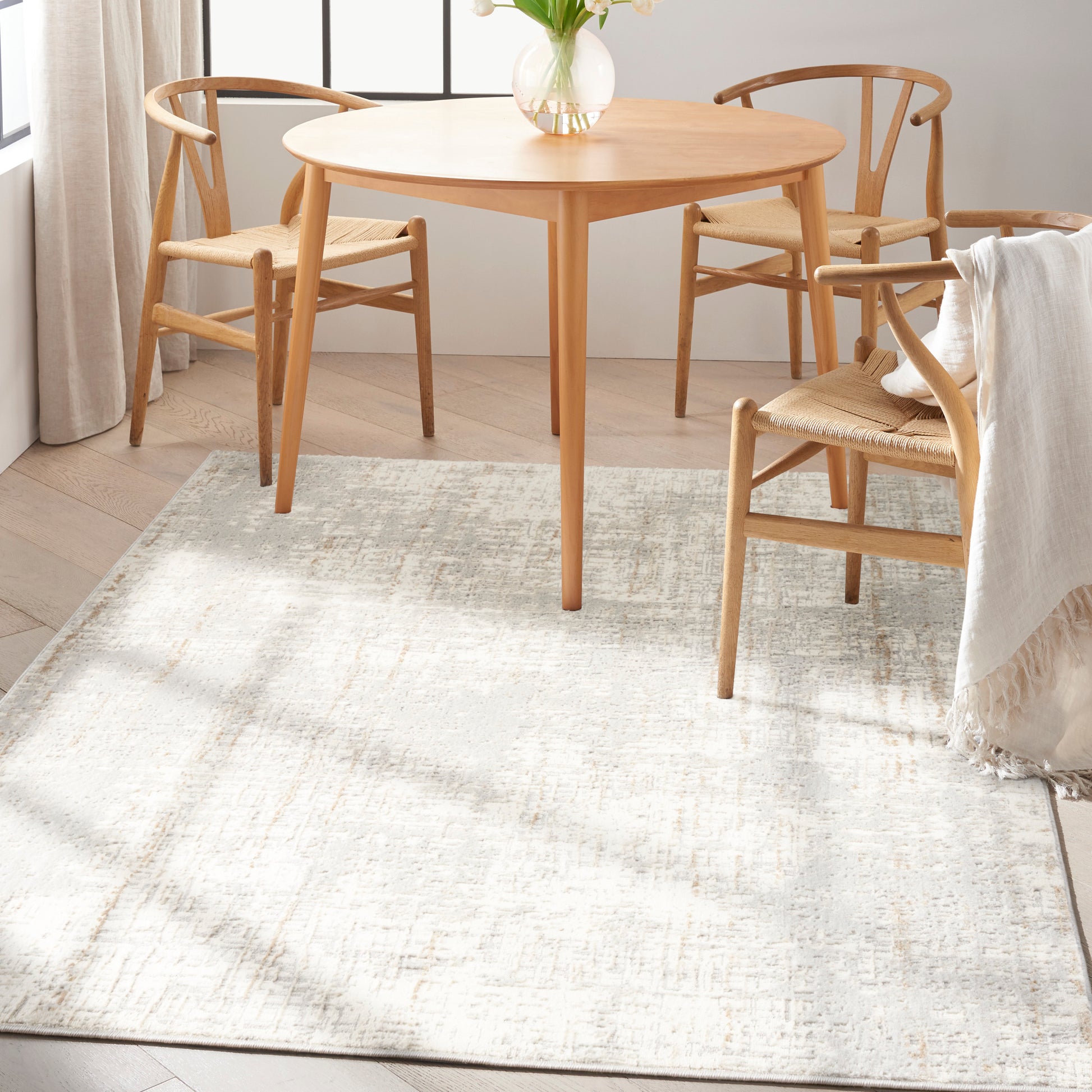 Round natural wood dining table with matching light oak chairs, woven seats, soft neutral rug and vase of flowers.