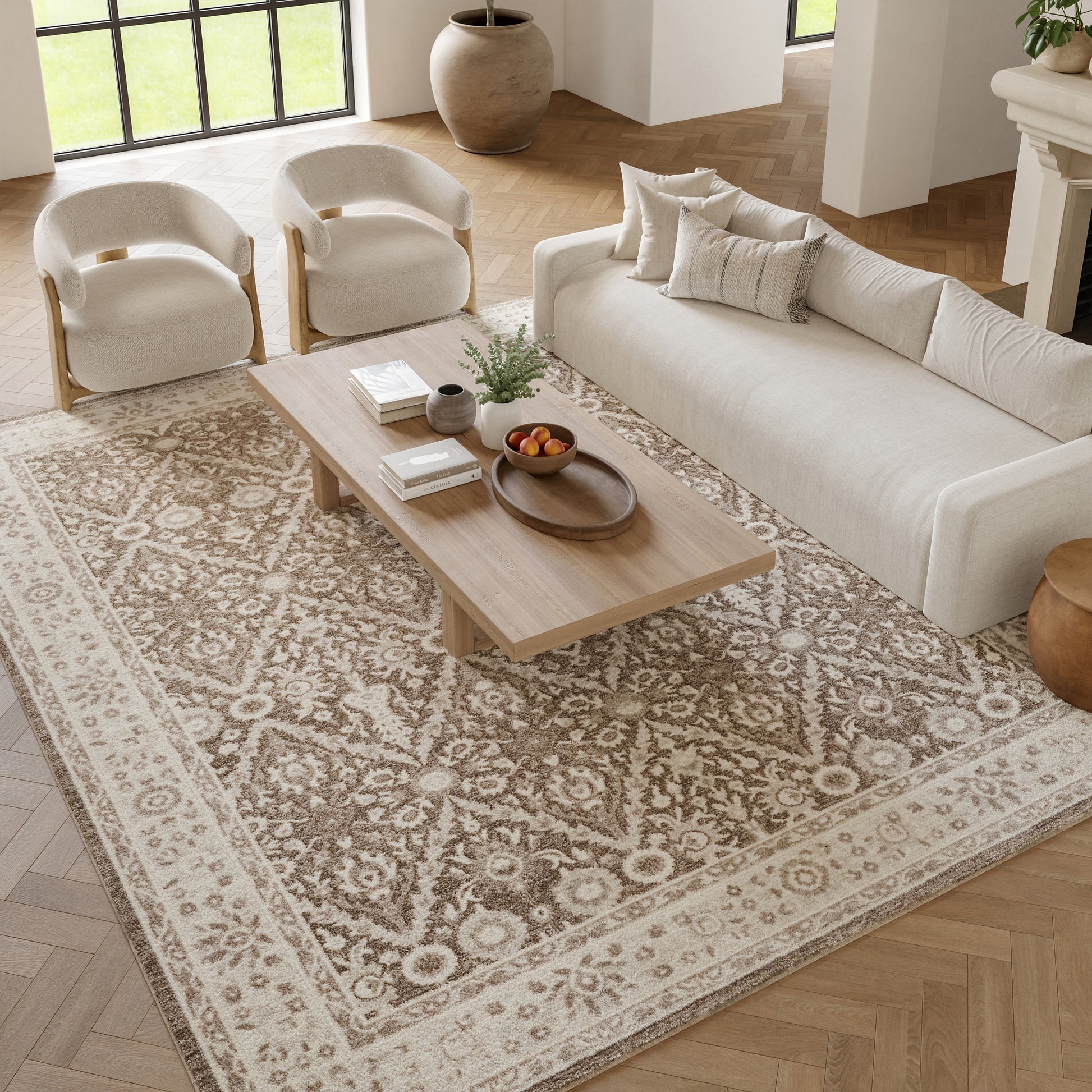 Neutral living room with cream sofa and two armchairs, wooden coffee table, beige ornate rug, plants and bowl.