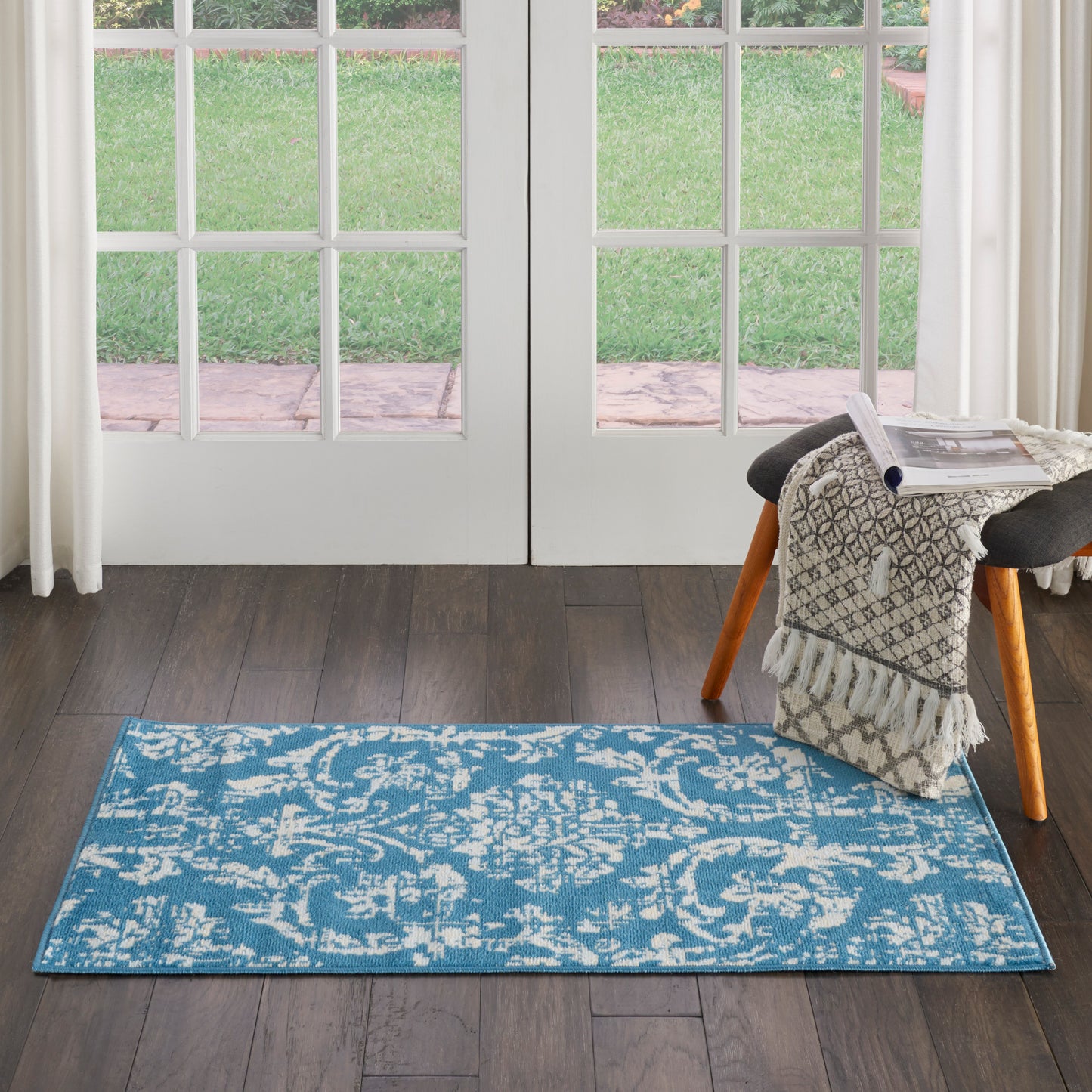 Blue ornate rectangular area rug on dark wood floor by glass French doors; fringed throw on a small stool.