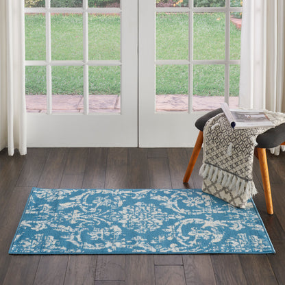 Blue ornate rectangular area rug on dark wood floor by glass French doors; fringed throw on a small stool.