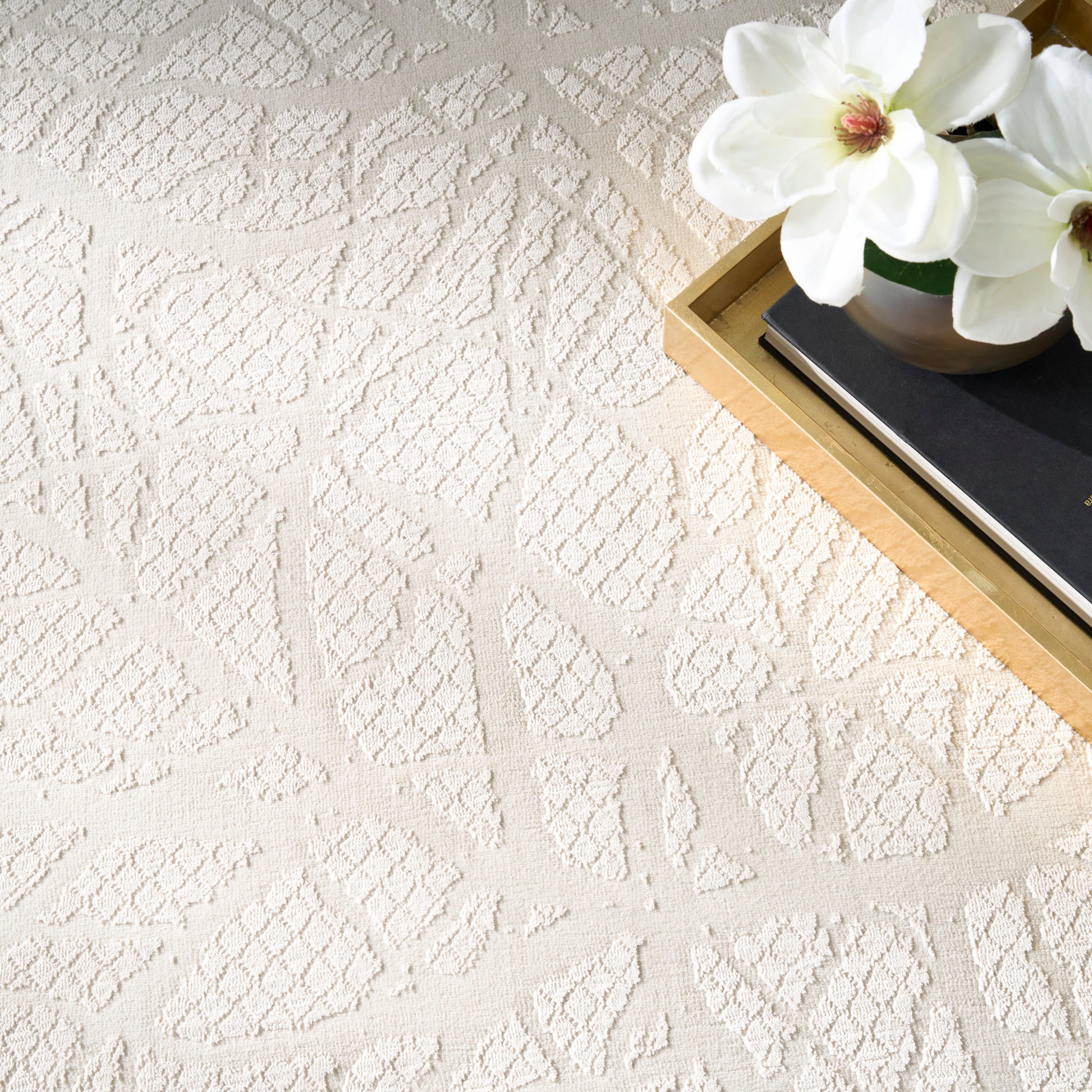 Cream textured geometric rug with diamond patterns; a gold-edged tray holds a black notebook & white magnolia flowers.