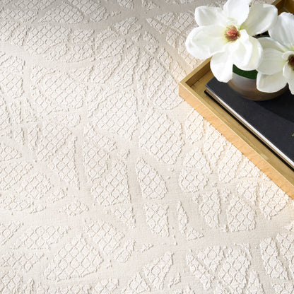 Cream textured geometric rug with diamond patterns; a gold-edged tray holds a black notebook & white magnolia flowers.