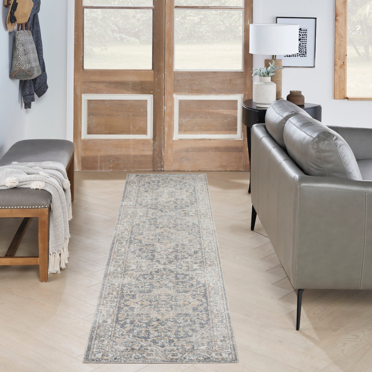Gray-blue ornate runner rug in a bright entryway with a beige bench, gray sofa, wooden doors, and a side table lamp.