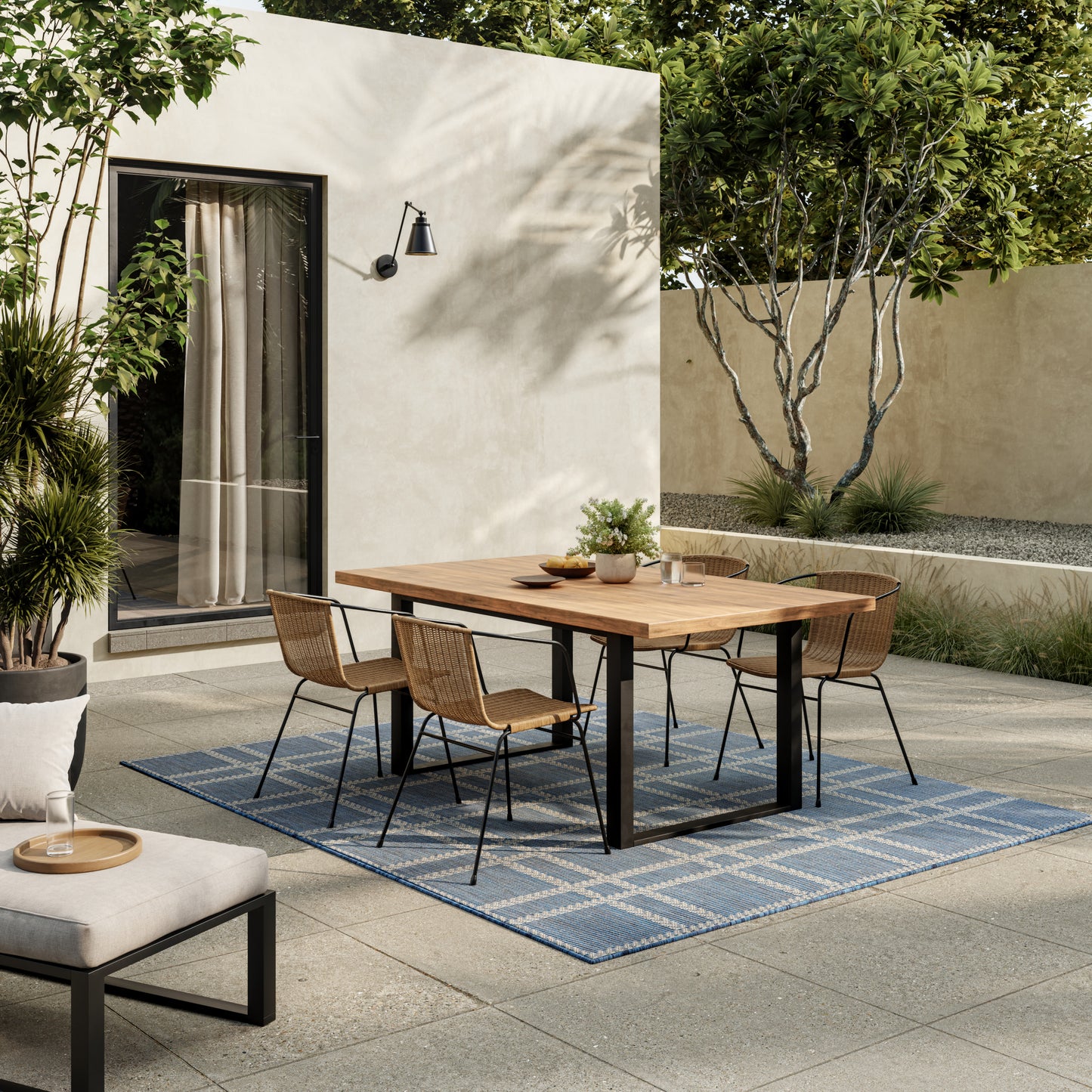 Outdoor dining set with wooden table and metal legs, wicker chairs, blue geometric rug, beige walls and lush greenery.