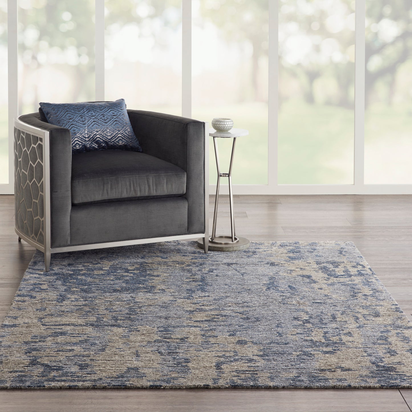 Dark gray velvet armchair with hexagonal metal frame, blue patterned pillow, glass-topped side table on a blue-gray rug.